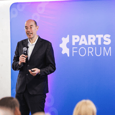 A man is standing in front of an audience. The background is purple and faded, it includes the PARTS FORUM logo in white. The man is called Danilo Zatta and is author and senior partner at implement.