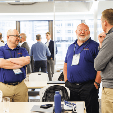 a group of men talking. Two wear a purple poloshirt with a logo on them and attendee badges aroundt heir necks. The man on the left wears glasses and is smiling. the man on his right is talking and has his hands in this black troursers pocks. The man on the right wears a grey shirt and beige pants during PARTS FORUM 2026 in Chicago.